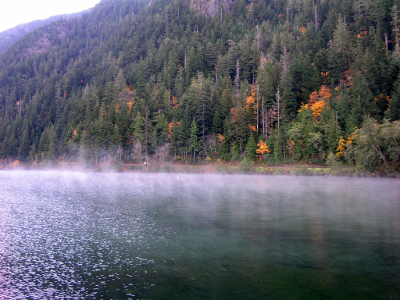 [A layer of mist rises in segments above the water. On the shore to the right is a high hillside a few trees with orange leaves or needles and all the rest of the trees are evergreens.]